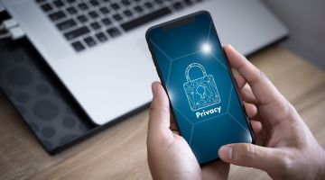 Smartphone in der Hand mit einem Privacy Schloss auf dem Bildschirm und einem Laptop im Hintergrund